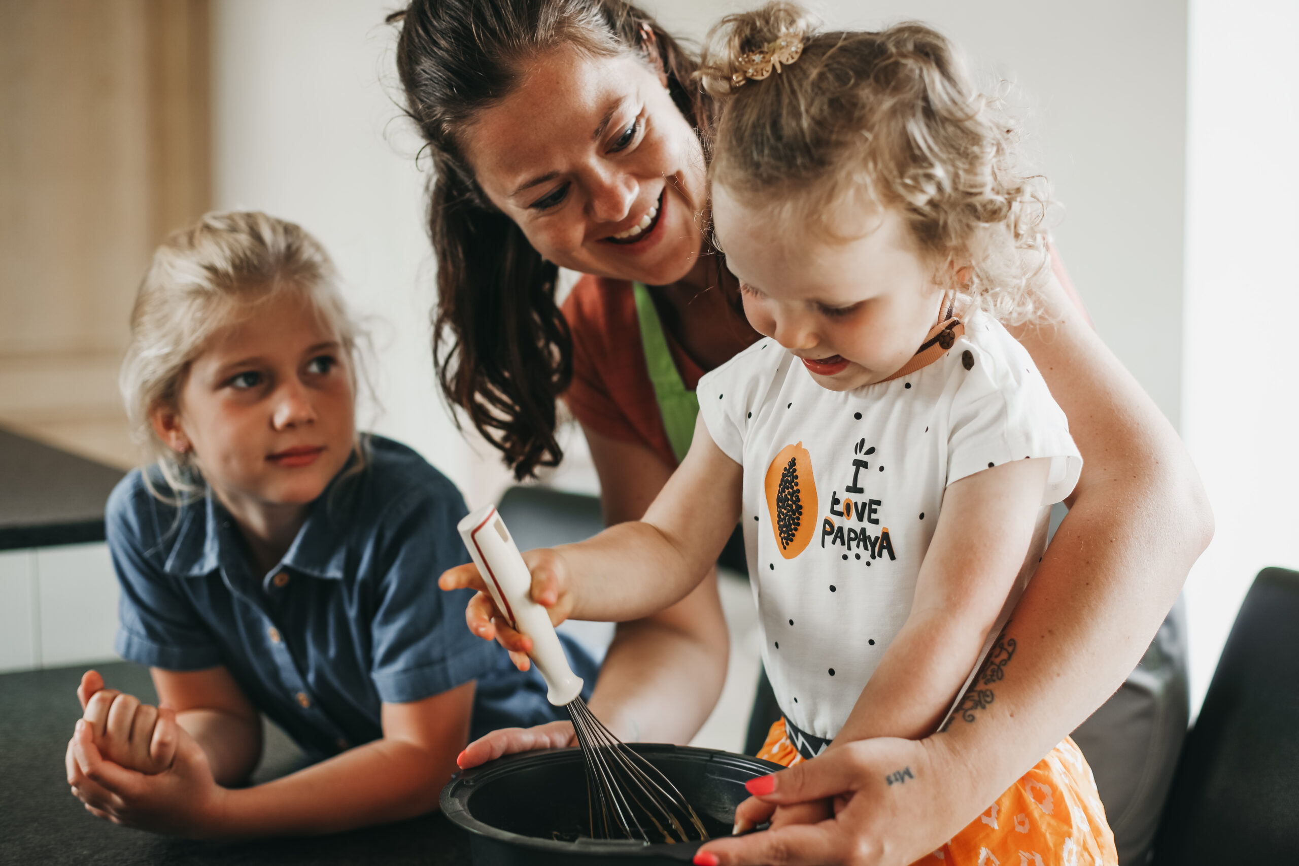 Een verzorgende van Familiezorg zorgt voor twee jonge meisjes. De verzorgende is aan het koken en laat een van de twee meisjes meehelpen door haar in de pot te laten roeren.