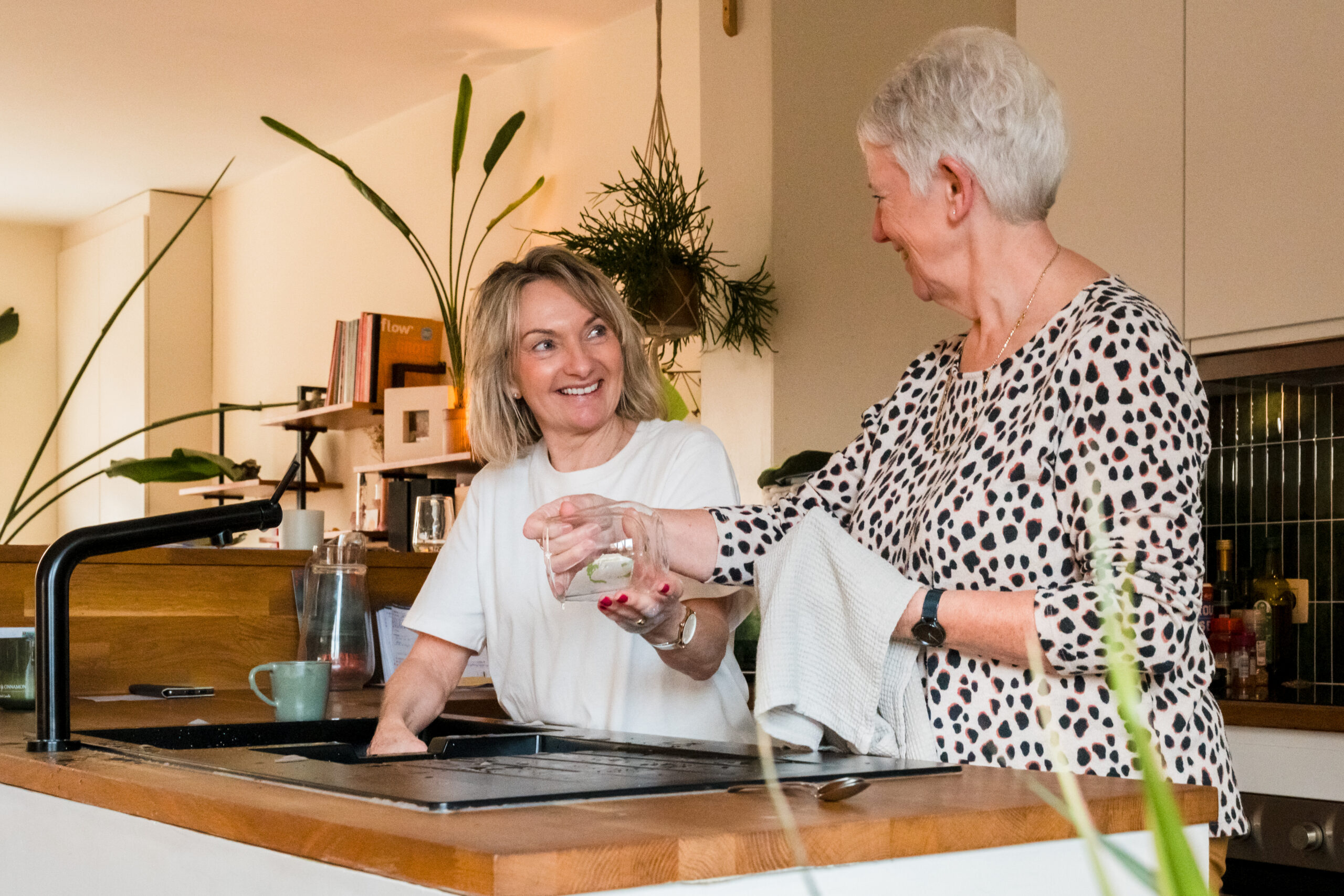 Een poetshulp van Familiezorg helpt een oudere vrouw, een cliënt van Familiezorg, met de afwas. De poetshulp wast een glas af en geeft ze door aan de cliënte om ze af te drogen.