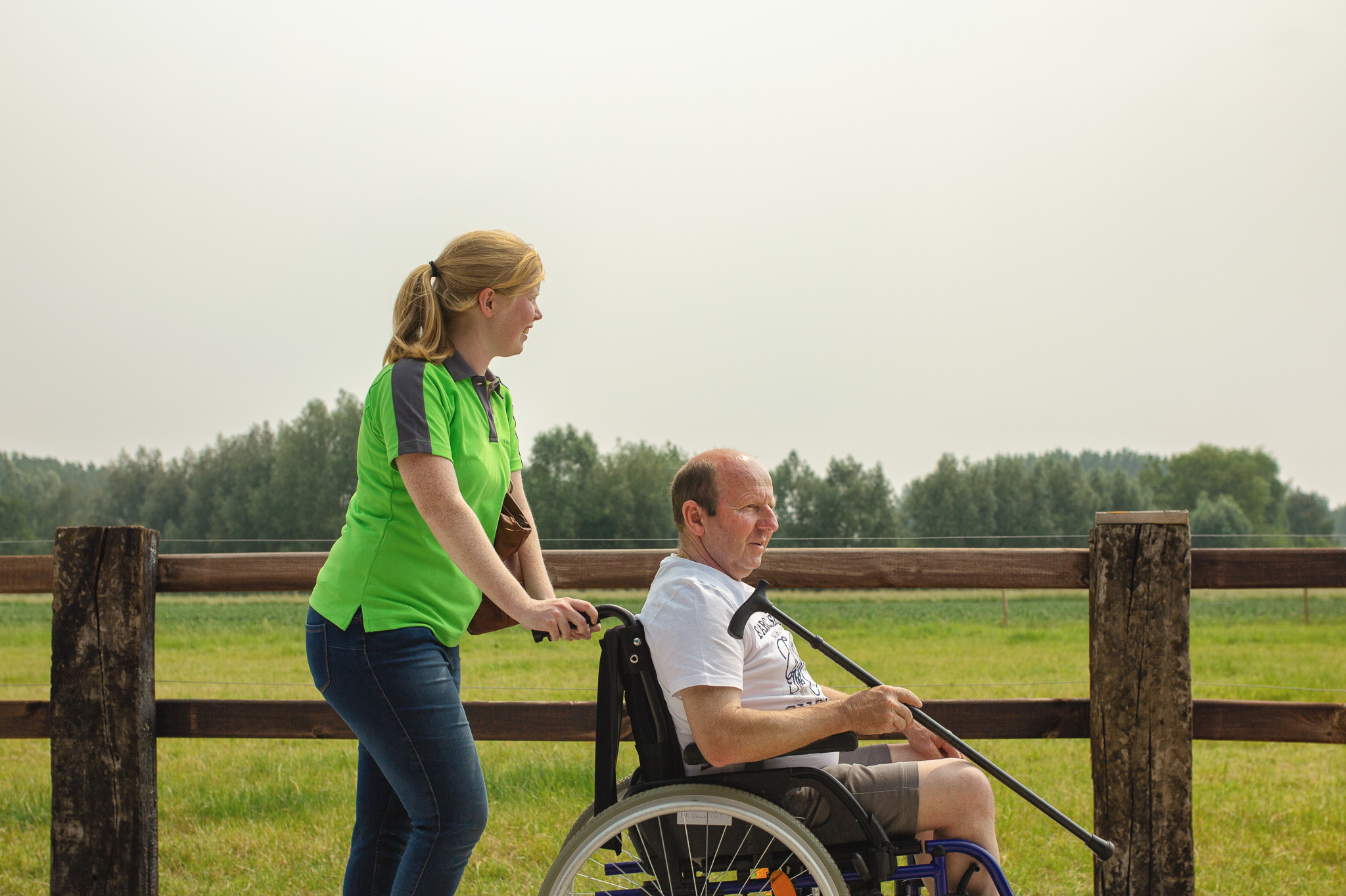 Een verzorgende van Familiezorg wandelt met een cliënt in een rolstoel langs een weide. De cliënt met jongdementie heeft ook een wandelstok in de hand.