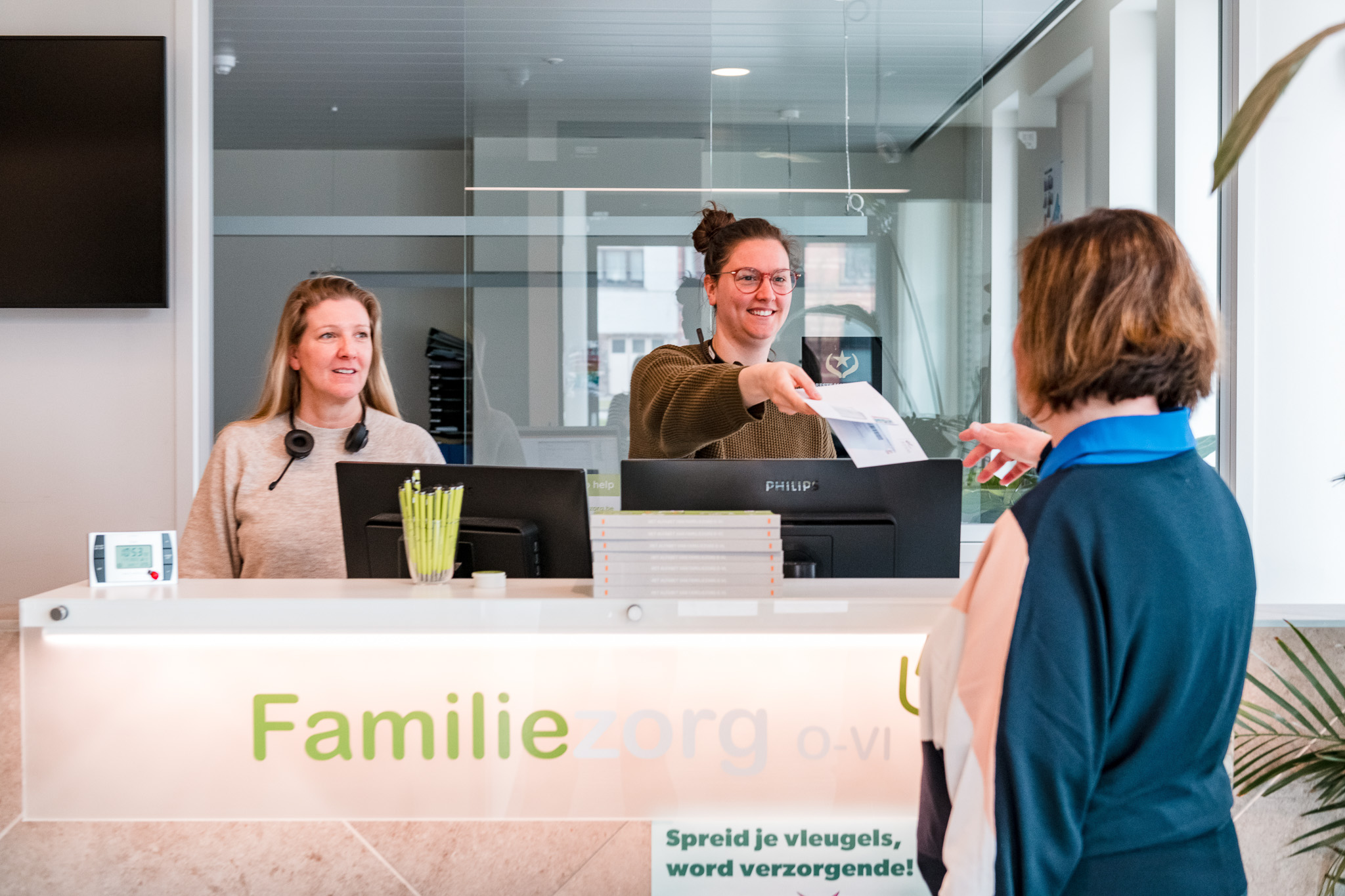 Twee onthaalmedewerkers van Familiezorg ontvangen een bezoeker aan de balie van het gebouw.