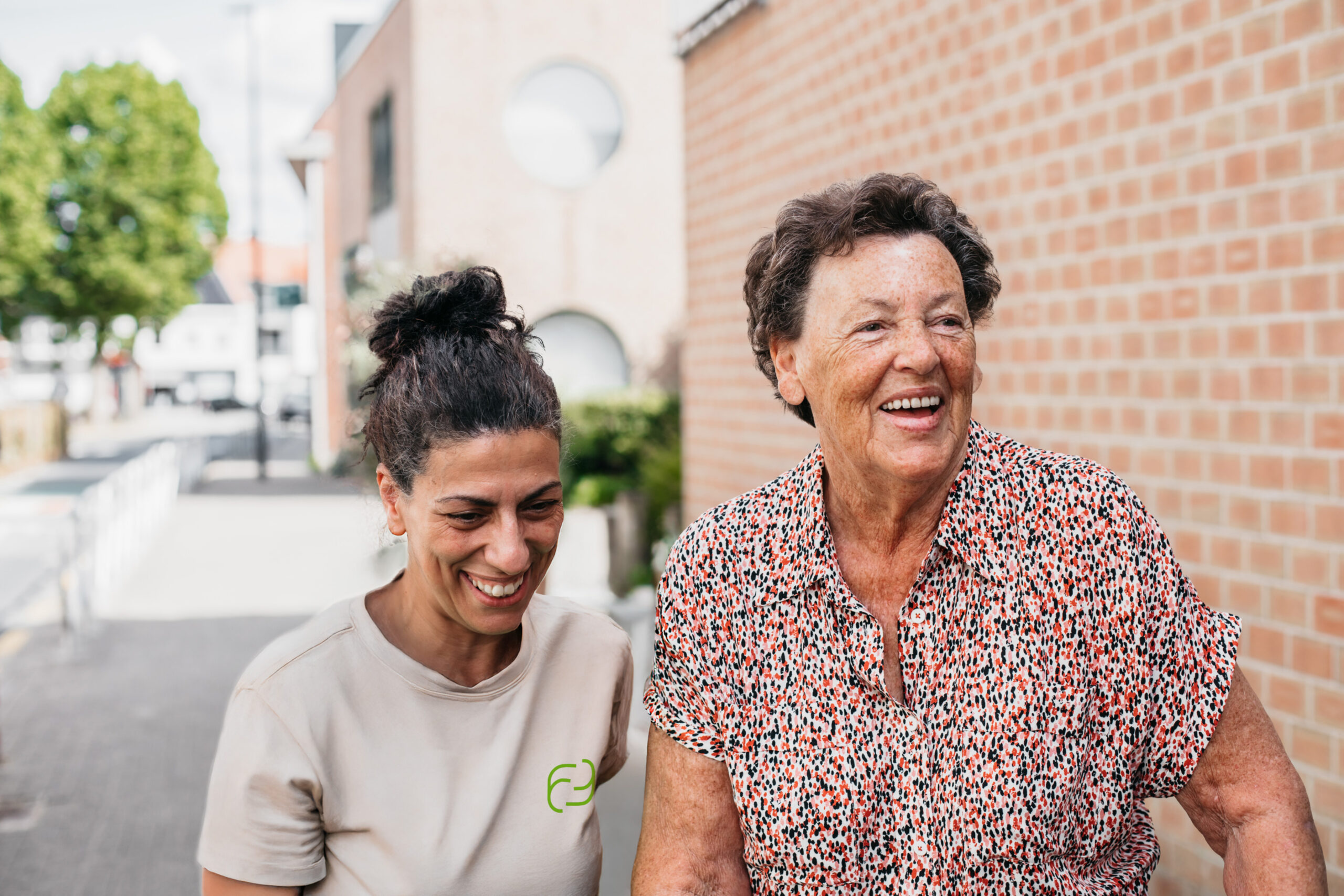 Verzorgende Familiezorg met beige t-shirt met groen logo. Oudere cliënt draagt kleurrijk kleed. Verzorgende en cliënt zijn blij en lachen.