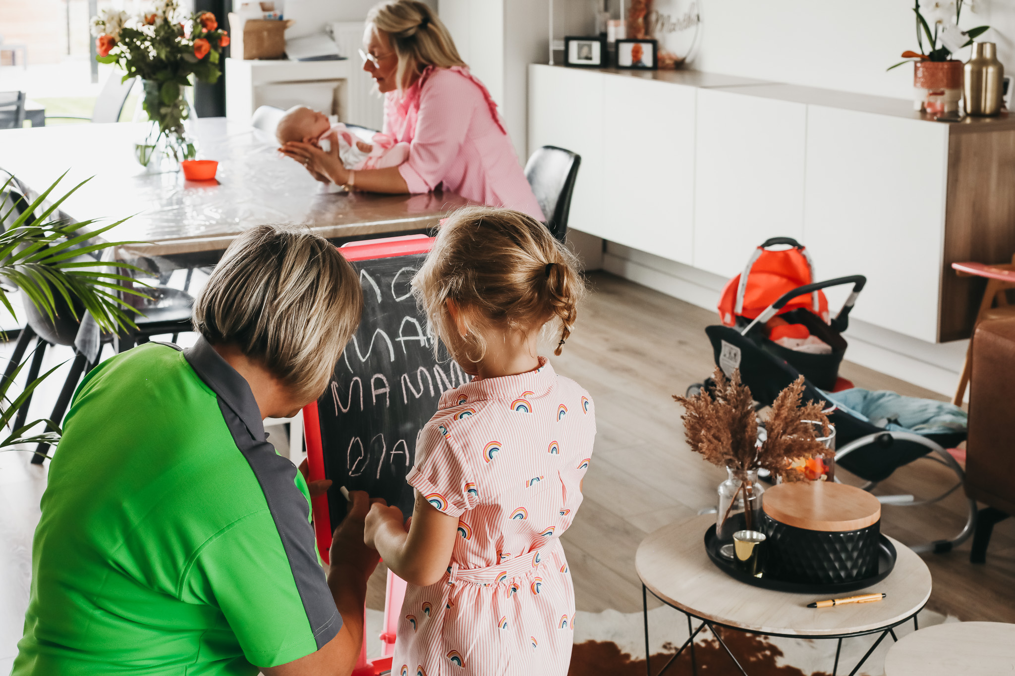 Kraamzorg verzorgende van Familiezorg die een meisje leert schrijven op een krijtbord. Ondertussen houdt de mama zich bezig met haar baby.