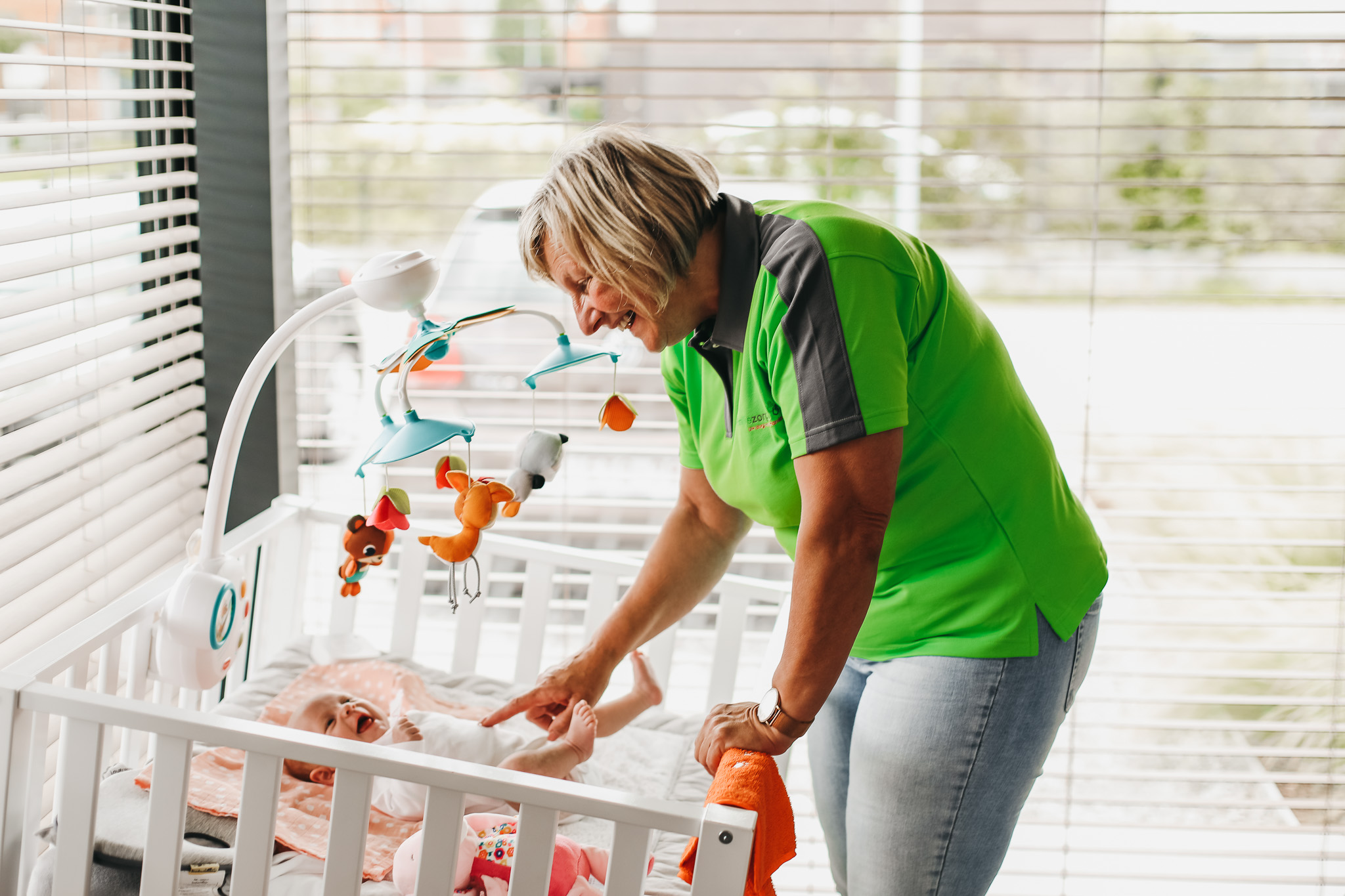 Kraamzorg verzorgende van Familiezorg die een baby in een wieg doet lachen. Aan de wieg hangt een mobiel met dierenknuffels.