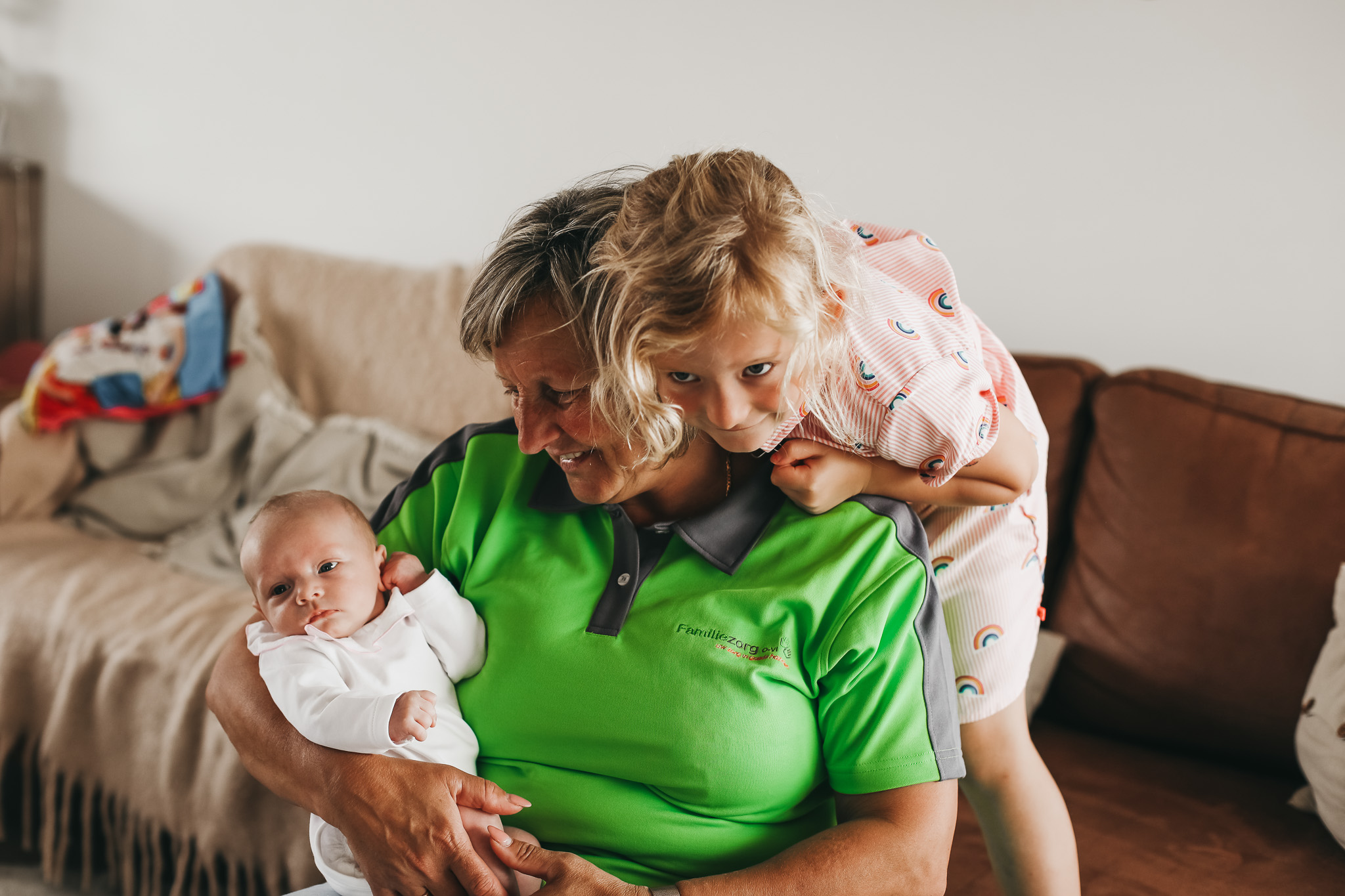 Kraamzorg verzorgende van Familiezorg met een baby in haar armen. Een ander meisje leunt op de schouder van de verzorgende. Ze zitten met drie in een zetel.