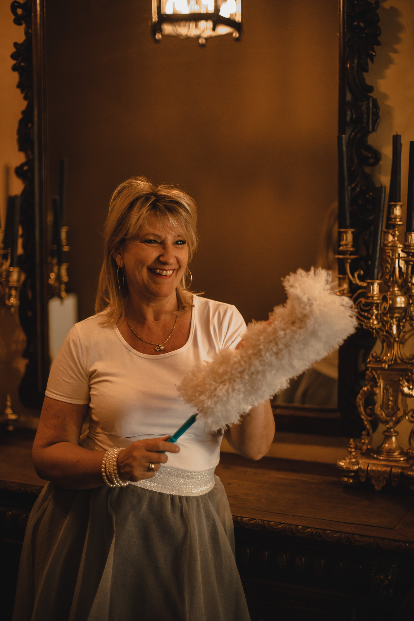 Poetshulp van Familiezorg met een witte pluizige plumeau in haar hand. Ze draagt een witte tshirt en staat naast een grote spiegel.